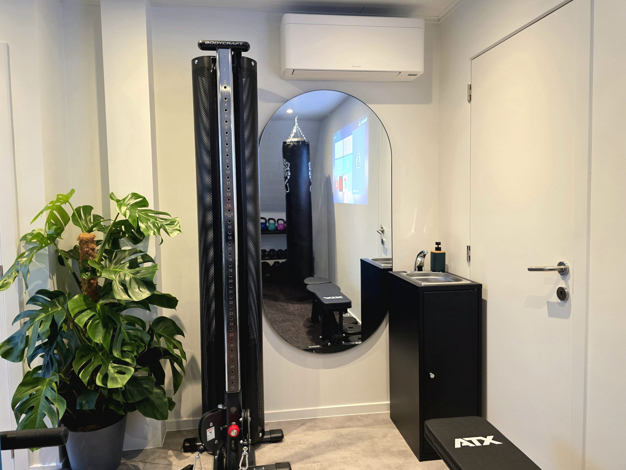 BodyCraft‑kabelmachine en bokszak Leo Punching Bag in moderne privé fitness Level170 met spiegel, groene plant, wastafeltje en projectie op de muur.