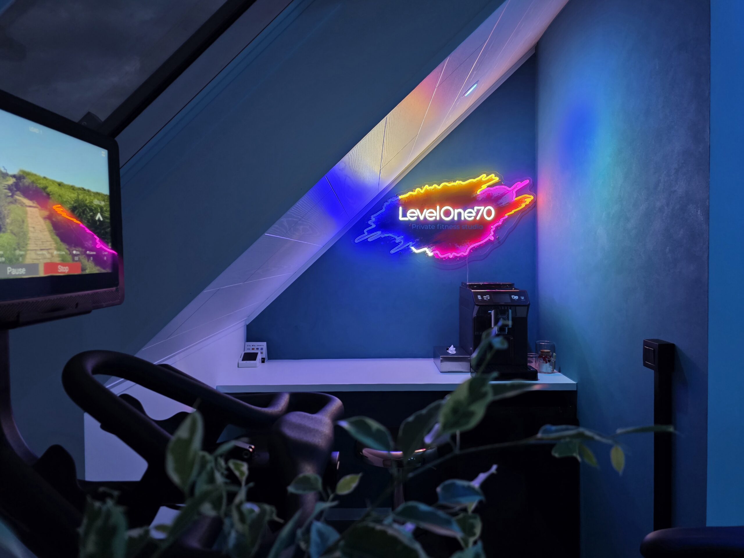 Fiets‑trainer met digitaal scherm en kleurrijk LevelOne70‑logo in privé fitness Level170, moderne trainingshoek met koffiemachine.